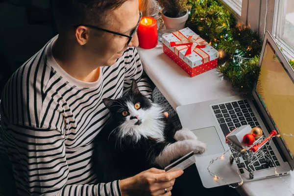 happy young man with funny cat holding credit debt card shopping online purchase xmas presents on laptop home. Christmas gift box, New Year winter sale discount. Safe ordering delivery at quarantine