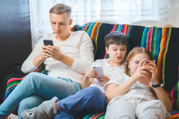 Cheerful young family dad son teen daughter children watching online video on smartphone sitting on couch together, laughing parent kids texting playing games entertaining using mobile phone apps home