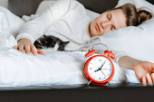 mladá tisíciletá upřímná žena spící s kočkou na bílé přikrývce útulná doma v ložnici. Close up of alarm clock, morning time, wake up, female healthy lifestyle concept