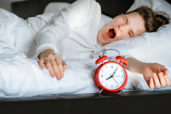 sleepy young millennial woman waking up in white bed yawning rubbing eyes turning off red vintage retro alarm clock in bedroom. Day healthy sleep. Relaxing concept. Candid moments in daily routine