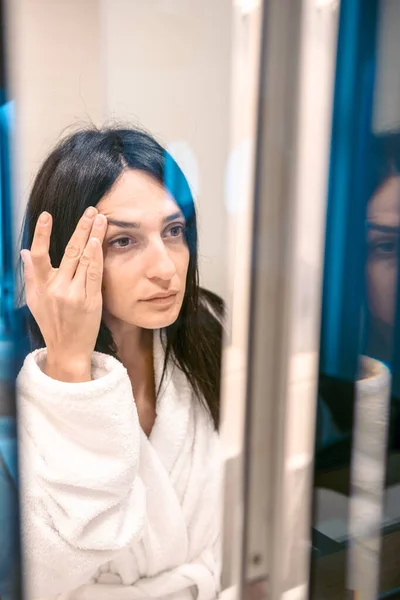 Young caucasian woman in a mirror touches with fingers skin above eyes.Aging and beauty procedures concept.