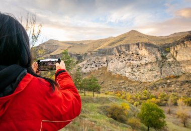 Arka taraftaki kadın dokunmatik ekranlı akıllı telefonu tutuyor ve Vardzia 'daki ünlü tatil yeri mağara şehrini yakalamak için basın mensupları ile konuşuyor. Georgia macerası.