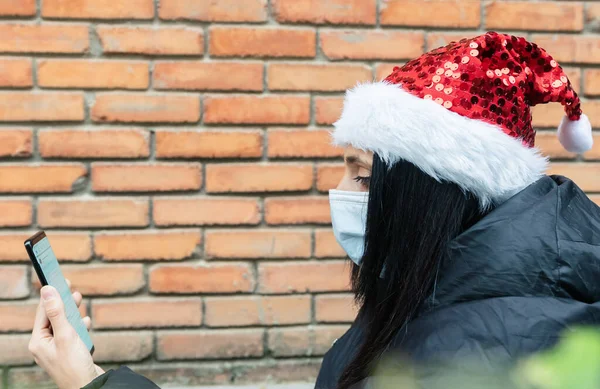 Young woman holds phone up and reads post on social media with xmas hat on and brick wall blank space backgroud. News media and social distancing duir corona virus pandemic.