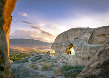 Uplistsikhe mağara şehir yapılarına, arka planda dramatik bir gökyüzü ve kayanın üzerinde duran turist manzarası.