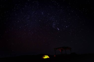 Karanlığın içinde parlak sarı bir çadır var. Kamp masasının arkasında, parlak mavi gökyüzü arka planında.