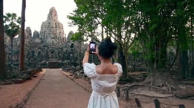 Beyaz elbiseli kadın Angkor Thom Kamboçya 'da orman ağaçlarıyla çevrili antik Bayon Tapınağı kalıntılarının akıllı telefon fotoğraflarını çekiyor. Seyahat ve tarih turizmi kavramı.