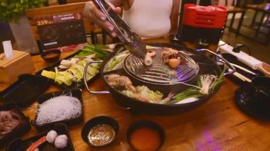 Bangkok, Tayland - 26 Şubat 2023: Bangkok 'ta Moo Krata olarak bilinen Tayland BBQ ızgarasında deniz ürünü ve sebze pişiren kadın. Masada karides, kalamar, lahana, mısır, mantar, erişte ve sos yer alıyor..