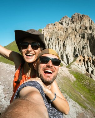Gülümseyen iki yürüyüşçü arkadaş dağın tepesinde selfie çekiyorlar. Milenyum erkeği kız yaz tatilinde kameraya gülerek eğleniyor Milenyum gezginleri manzaralı doğada duruyor.