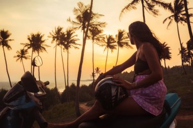 Romantik genç beyaz kadın kırmızı scooter 'da oturur günbatımını seyreder Asya' da tropikal yalnızlığın tadını çıkarır açık havada tropikal palmiyelerle çevrili