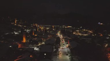 Night aerial view forwards of illuminated Mestia mountain ski resort village in winter with holiday decorations, snowy rooftops, and festive street ambiance