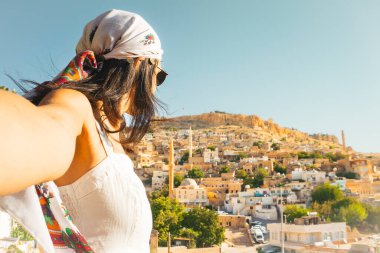 Güneş gözlüklü gülümseyen kadın, tarihi Mardin Türkiye 'nin panoramik silueti ve Mezopotamya yamaçlarındaki cami minareleri ile selfie çekiyor.