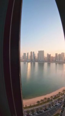 Şafak vakti, balkon penceresi manzaralı, Sharjah rıhtımına bakan, sakin deniz ve plaj, gündoğumundaki kentsel büyüme ve kıyı emlak yatırımlarını vurguluyor.