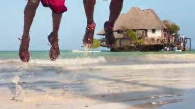İki Masai adamı Zanzibar plajında geleneksel atlayış yapıyorlar. Arka planda ikonik rock restoranı ve turkuaz Hint Okyanusu var. Kültürel seyahat kavramı.