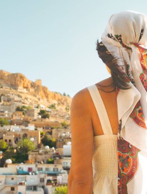 Beyaz elbiseli gezgin kadın tarihi Mardin şehrinin panoramik manzarasına bakıyor cami minareleri ve geleneksel evleri ve tepedeki Mardin şatosuyla