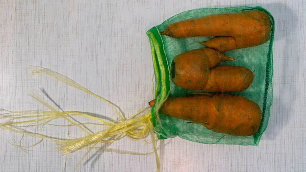 Three ugly carrots in green mesh bag lie on the striped rustic table. Farmers vegetables is good for nutrition. It\'s contains vitamins and microelements. Top view horizontal image with copy space.