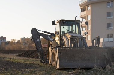 Tamamlanmış dairelerin önünde kepçe doldurucu. Kazıcı gün batımında bir hendeğin yanına park etmiş. İnşaat alanı. Yeni daireler. Emlak piyasası