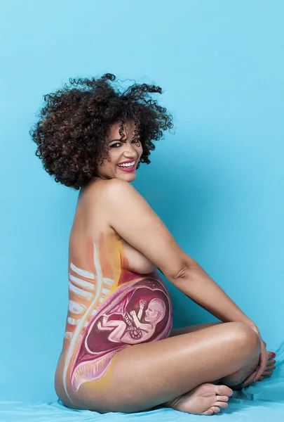 Cheerful pregnant woman sitting with baby painted on belly — Stock Photo, Image