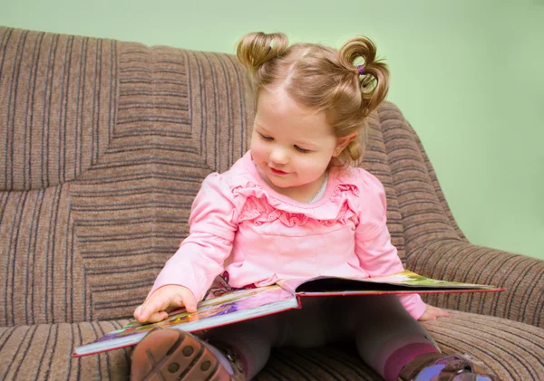 Söt liten flicka sitter på en soffa och tittar på en bild boo Stockbild