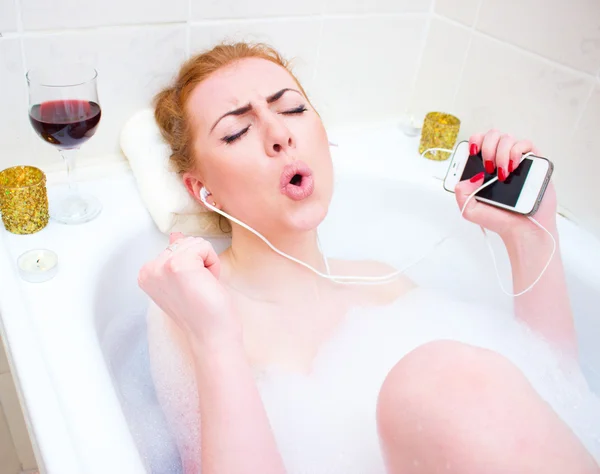 Young woman  listening music while lying in the bath with foam and candles — Zdjęcie stockowe