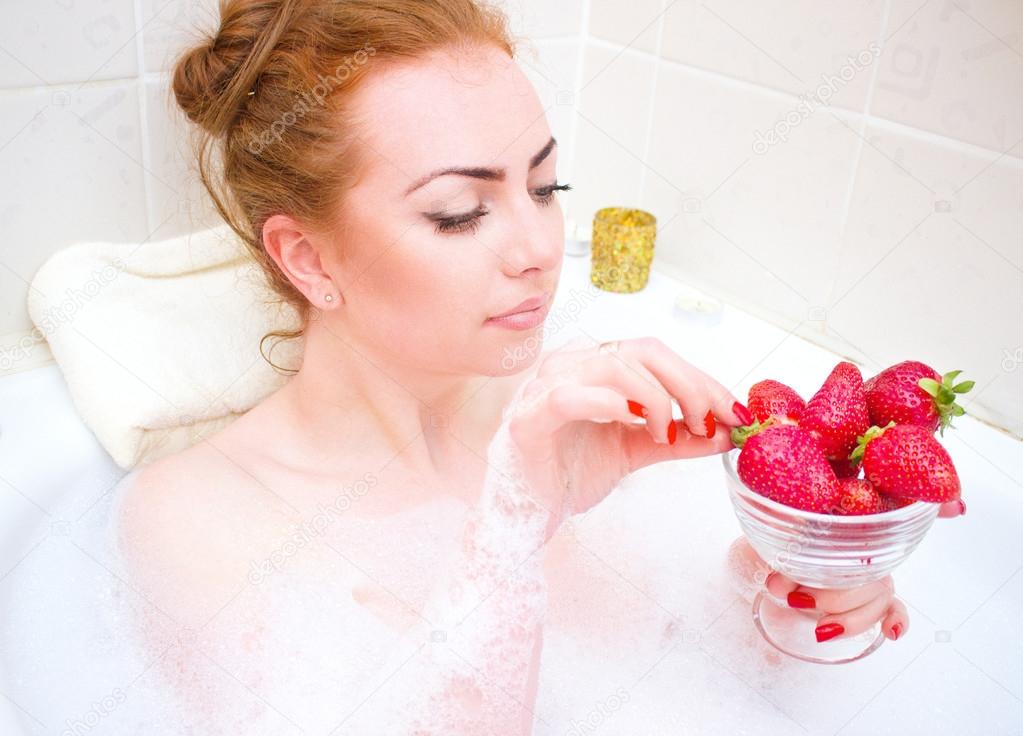 Young woman  eating strawberry while lying in the bath with foam and candles