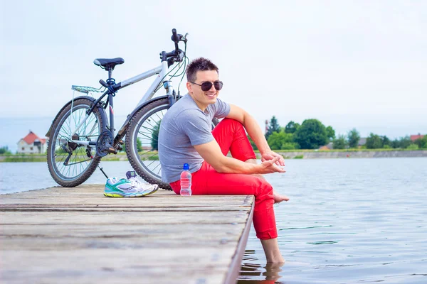 Junger Mann, ein fröhlicher Tourist mit grauem Hemd und Brille sitzt am Strand und ruht sich nach der Radtour aus. — Stockfoto