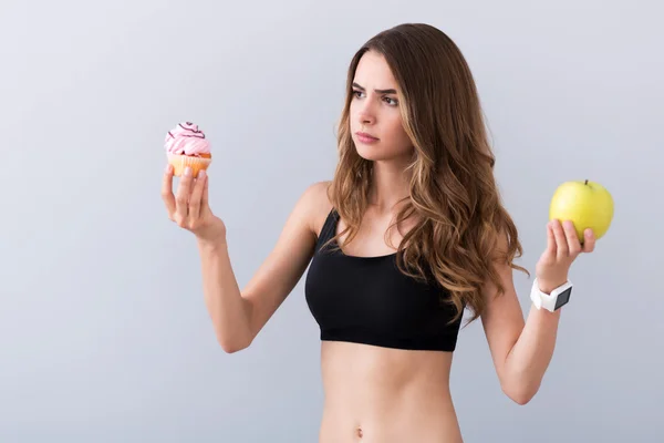 Piacevole donna perplessa che tiene frutta e cupcake — Foto Stock