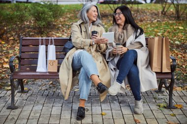 Neşeli kadınlar bankta oturup tablet bilgisayar kullanıyorlar.