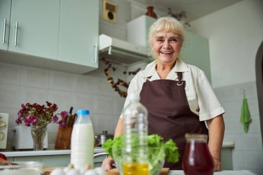 Önlüklü neşeli yaşlı kadın mutfakta duruyor.