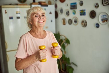 Neşeli yaşlı kadın evde dambıllarla egzersiz yapıyor.