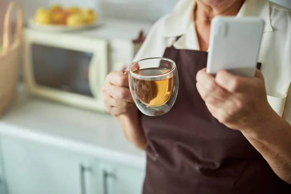 Starsza kobieta trzyma filiżankę herbaty i telefon komórkowy — Zdjęcie stockowe