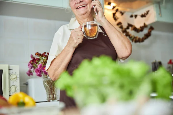 Gioiosa vecchia che parla al cellulare e beve tè a casa — Foto Stock