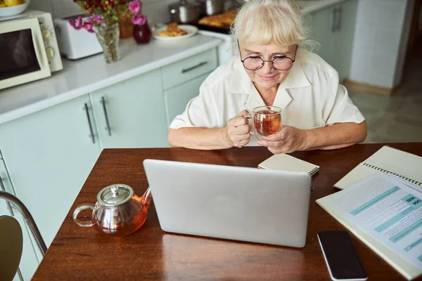 Schöne alte Frau trinkt Tee und benutzt Laptop zu Hause — Stockfoto