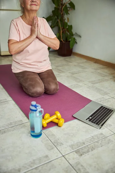 Dizüstü bilgisayar kullanan ve evde yoga yapan sakin yaşlı bir kadın.