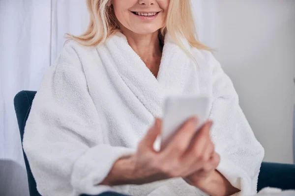 在温泉中心，穿着白色长毛绒浴衣的快乐微笑的女人使用手机和互联网 — 图库照片