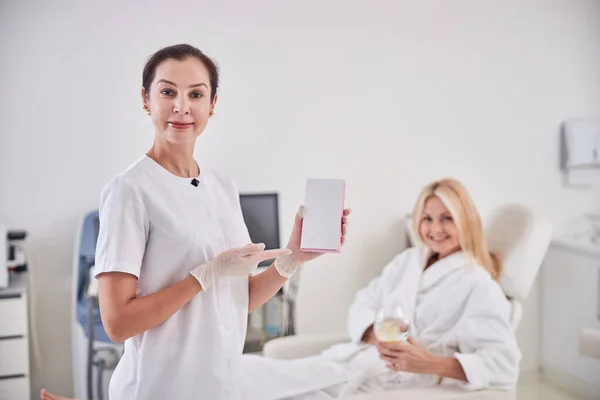 Hermoso cosmetólogo profesional de pie cerca del paciente con cuaderno blanco en la habitación en el interior — Foto de Stock