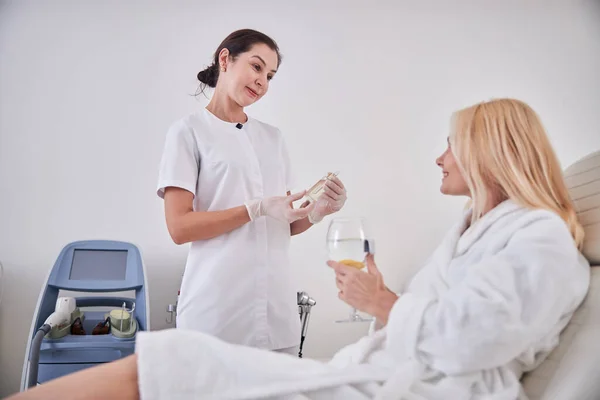Albornoz blanco mujer rubia feliz sosteniendo vaso de agua mientras escucha a su cosmetólogo en la habitación en el interior — Foto de Stock