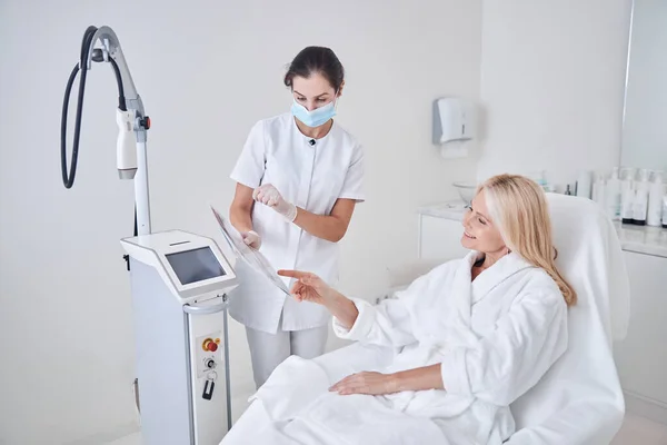 Happy smiling client looking to the list with programs of ultrasound modern machine in beauty center — Stock Photo, Image