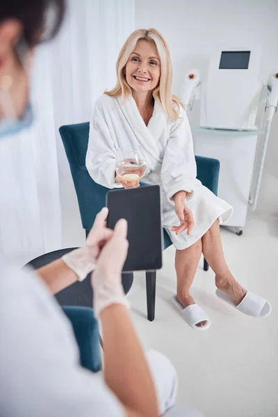 Hermosa hembra sosteniendo vidrio con agua en la mano mientras habla con el médico en el gabinete — Foto de Stock