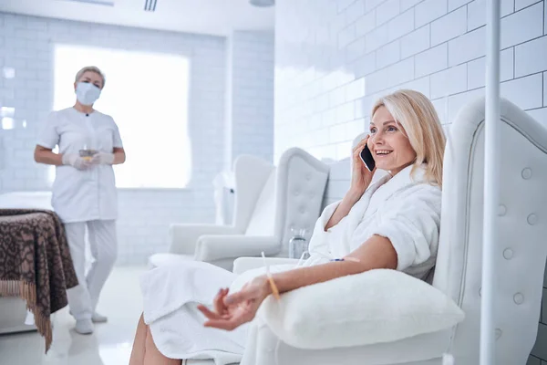 Glücklich lächelnde blonde Frau im weißen Bademantel sitzt im Schönheitsstuhl bei der Behandlung Vitamintropfen — Stockfoto