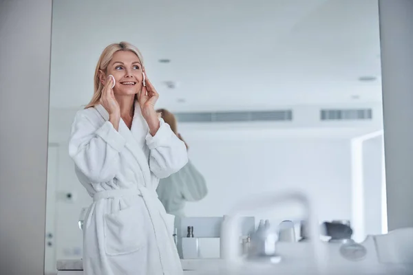 Gyönyörű felnőtt nő fehér plüss fürdőköpenyben takarítás bőr arc a fürdőszobában — Stock Fotó