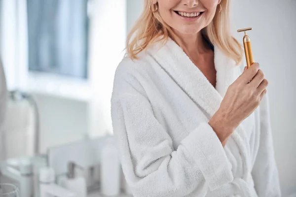 Indah menarik perempuan dalam mantel mandi mewah putih menggunakan rol wajah saat berdiri di dalam ruangan — Stok Foto