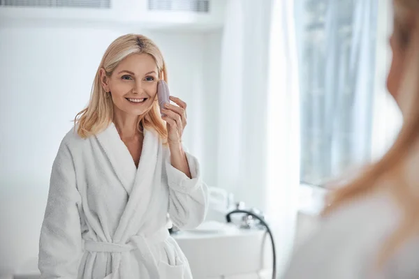 Boldog mosolygós nő fehér fürdőköpenyben nézi a tükröt a fürdőszobában — Stock Fotó
