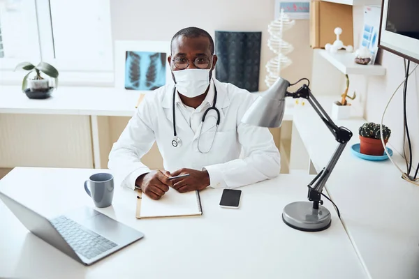 Médico com uma máscara posando para câmera em sua mesa — Fotografia de Stock