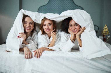 Üç kadın beyaz çarşaflarla yatağında pijama partisi veriyor.