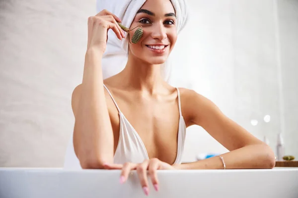 Joyous signora in una vasca da bagno massaggiare il suo viso con rullo — Foto Stock