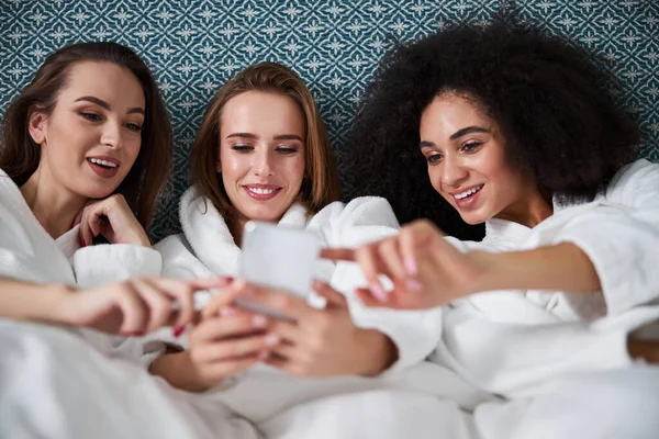 Cheerful girlfriends scrolling on smartphone while resting in bed — Stock Photo, Image