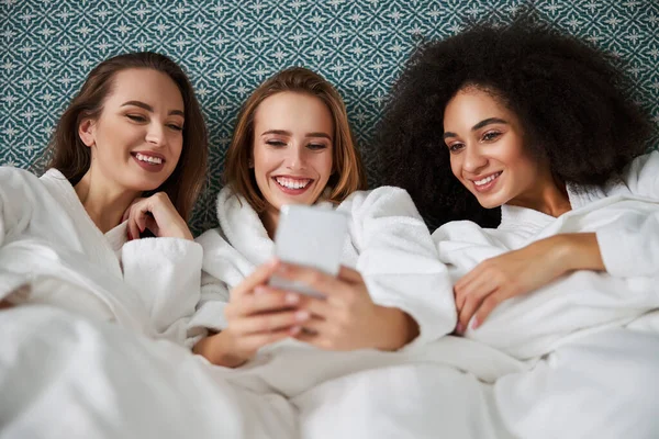 Senhoras alegres relaxando na cama e olhando para a tela do smartphone — Fotografia de Stock