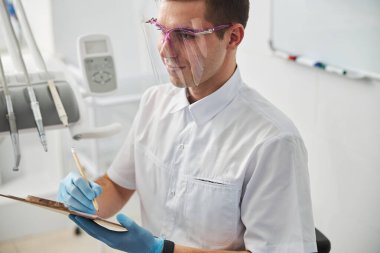 Yüz kalkanındaki doktor kağıda not alıyor.