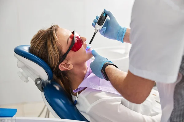 Dentista calificado tratando a hembra en gafas de seguridad con lámpara de fotopolímero — Foto de Stock