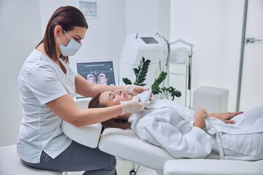 Beyaz bornozlu genç bayan güzellik kliniğinde profesyonel ultrason yüz gerdirme ameliyatı oluyor.